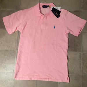 Rosa pikétröja från Ralph Lauren - Snygg ljusrosa pikétröja från Ralph Lauren med klassisk krage och korta ärmar. Tröjan har blå broderad logga på bröstet och knäppning med två knappar. Tillverkad i mjuk bomull som känns skön mot huden. Perfekt för en fräsch och stilren look.