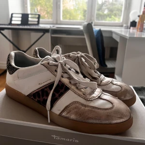 Tamaris sneakers med leopard och glitter - Snygga vita sneakers från Tamaris med metallic-detaljer, leopardmönstrade paneler på sidorna och glittrig häl. Skorna har vit snörning och en klassisk gummisula. Perfekt för dig som vill sticka ut med coola detaljer och djurmönster.