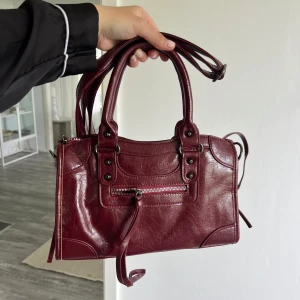 Vinröd handväska med dragkedja - Vinröd handväska i skinnimitation med coola metalldetaljer och dragkedja framtill. Väskan har både handtag och lång axelrem, så du kan bära den på flera sätt. Snygg design med dekorativa sömmar och detaljer som ger en edgy vibe.
