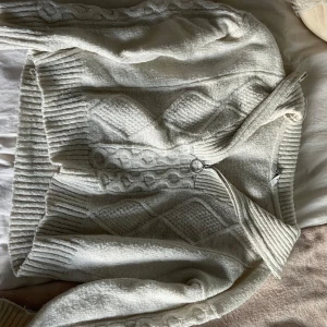 Vit stickad kofta med dragkedja - Supermysig vit stickad kofta med snyggt kabelstickat mönster och ribbade muddar. Koftan har en cool dragkedja med ring och en klassisk krage. Perfekt för lager-på-lager och chill dagar.