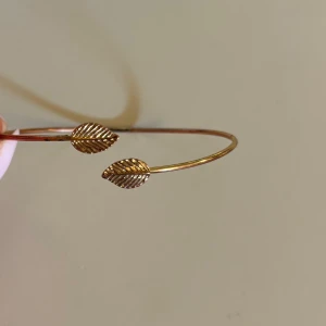 Guldigt armband med lövdetaljer - Snyggt och minimalistiskt armband i guldton med två detaljerade löv i ändarna. Armbandet är tunt och öppet, vilket gör det enkelt att ta på och av. Perfekt accessoar för att ge din outfit en naturell och trendig touch. Bra skick. Onesize