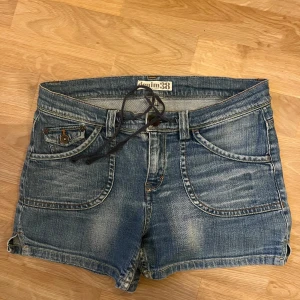 Lowaist jeansshorts med snörning - Snygga blå jeansshorts från Denim38 med cool snörning i midjan och slitna detaljer. Klassiska fickor fram och bak med knappar och kontrastsömmar. Perfekta för sommaren och ger en avslappnad vibe till din outfit.