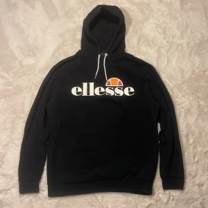 Svart Ellesse hoodie  - Svart hoodie från Ellesse i storlek M med stor vit logga och orange detalj på bröstet. Tröjan har huva med vita snören och en klassisk känguruficka framtill. 