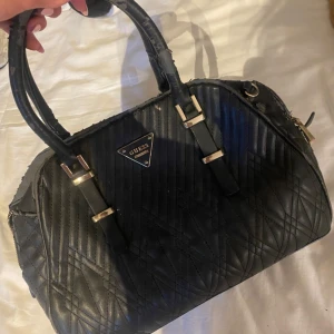 Svart quiltad handväska från Guess - Snygg svart handväska från Guess med quiltad yta och guldfärgade detaljer. Väskan har dubbla handtag, dragkedja och ett coolt leopardmönstrat innerfoder. Perfekt storlek för allt du behöver ha med dig och ger en lyxig vibe till din outfit. Den har en del tecken på användning då den börjat ”flagna” lite på vissa ställen vilket du kan se på bilden.