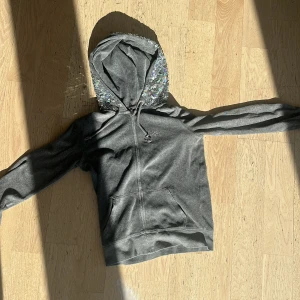 Grå hoodie med paljetter från PINK - Grå hoodie från PINK by Victoria's Secret i storlek XS. Huvan och texten på ryggen är täckta av glittriga paljetter. Tröjan har dragkedja framtill, känguruficka och snörning i huvan. Perfekt för dig som gillar lite extra bling i vardagen.