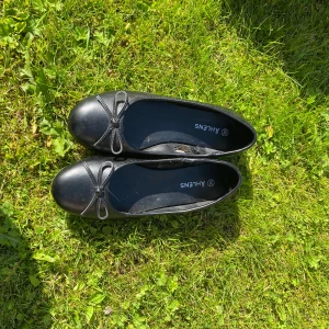 Svarta ballerinaskor - Klassiska svarta ballerinaskor från Åhléns med rund tå och söt rosettdetalj framtill. Skorna har platt sula och är tillverkade i ett syntetmaterial som ger en stilren look. Perfekta att matcha med jeans eller klänning. Använda en gång.
