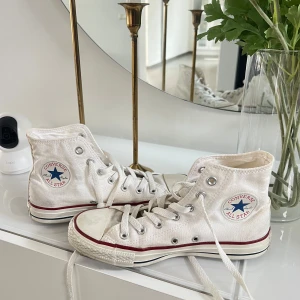 Vita Converse - Klassiska vita Converse All Star high tops med röd och blå rand längs sulan. Skorna har snörning, rund tå och är tillverkade i canvas med gummisula. Perfekta för en avslappnad och cool streetstil.