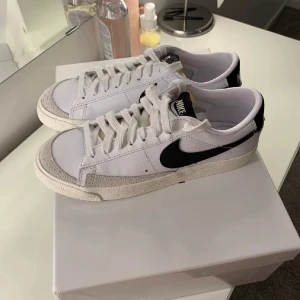 Nike Blazer Low vita sneakers - Snygga Nike Blazer Low sneakers i vitt med svart swoosh. Klassisk retrostil med lågt skaft, snörning och tåparti i mocka. Ovandel i textil och skinn, perfekt till streetwear eller casual outfits. Tidlös design som passar till det mesta.