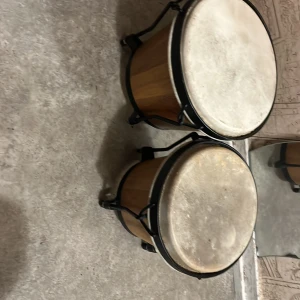 Bongotrumor - Bilderna visar ett par bongotrummor i trä med naturfärgade skinn och svarta metallspännen. Perfekt för dig som vill jamma, skapa beats eller bara ha kul med musik! Trummorna har en cool vintage-look och passar både nybörjare och erfarna musiker.