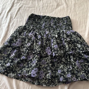 Svart blommig volangkjol - Supersöt kort kjol med svart botten och lila blommor. Kjolen har smock i midjan och dubbla volanger. Perfekt för dig som gillar blommigt och vill ha något bekvämt och snyggt till sommaren. Skulle säga att den är stor i storlek och går ca till halva låret.