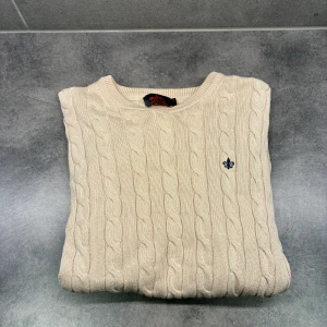 Morris tröja  - Kabelstickad beige tröja från Morris i 100% pima bomull. Klassisk rund halsringning och diskret broderad logga på bröstet. Perfekt för dig som gillar stilrena och tidlösa plagg med snygg struktur. Långärmad och normal passform.