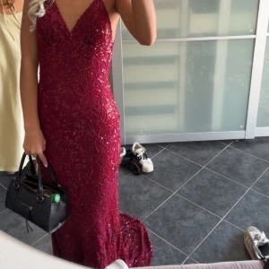 Vinröd glittrig långklänning med öppen rygg - Säljer en vinröd långklänning med glittriga paljetter och v-ringning. Klänningen har en figurnära passform, öppen rygg och smala axelband. Perfekt för bal eller fest och ger en riktigt glam känsla.