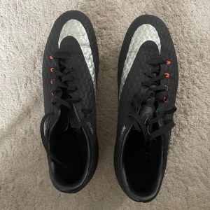 Nike svarta fotbollsskor strl 37,5 - Svarta Nike fotbollsskor med stora silvriga swoosh-loggor och orange detaljer vid snörningen. Skorna har plastsula med orange dobbar för bra grepp på planen. Lätta och smidiga, perfekta för dig som vill ha stilrena och snabba skor på matchen. Storleken är 37,5 passar även 37