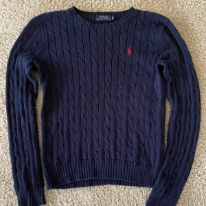 Mörkblå kabelstickad tröja Polo Ralph Lauren - Kabelstickad mörkblå tröja från Polo Ralph Lauren med klassisk rund hals och röd broderad logga på bröstet. Tillverkad i mjuk bomull och har långa ärmar samt ribbade muddar för en snygg passform.