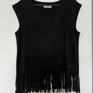 Svart topp, Nümph, storlek M - Svart ärmlös topp med fransar nedtill från Nümph i storlek M. Toppen har en rund halsringning och en liten dekorativ detalj på sidan. Mockaimmitation liknande material, 90% polyester och 10% elastan.  Nyskick.   Mått: Längd inklusive fransar ca 66 cm Byst ca 51 cm Midja ca 51 cm
