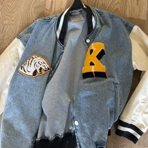 Jeans bomberjacka från Bershka - Säljer en snygg bomberjacka från Bershka i jeansmaterial med vita ärmar och coola patches, bland annat en tiger och ett stort gult K. Jackan har svarta och vita muddar och knäppning framtill. Perfekt för dig som vill sticka ut med en sportig och trendig look.