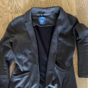 Mörkgrå kavaj från Tom Tailor - Snygg mörkgrå kavaj från Tom Tailor i klassisk modell med två fickor framtill och smal krage. Perfekt för dig som gillar stilrena plagg med enkel design. Materialet känns mjukt och följsamt, vilket gör den bekväm att bära hela dagen.
