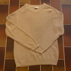 Beige stickad tröja från NA-KD - En stilren beige stickad tröja från NA-KD Reborn i storlek S. Tröjan har rund halsringning, ribbade muddar och är långärmad. Perfekt för lager-på-lager och enkel att matcha med jeans eller kjol. Snygg basic som funkar året runt.