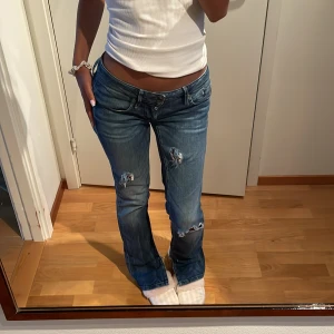 Lågmidjade bootcut jeans från G-Star - Säljer ett par blå lågmidjade jeans från G-Star med bootcut passform. Jeansen har slitna detaljer och hål på benen för en cool, avslappnad look. Klassisk femficksmodell med broderad G-Star-logga vid fickan. 
