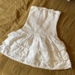 Vit axelbandslös volangklänning - Supersöt vit axelbandslös klänning från Gina Tricot med smockad överdel och två lager volanger i kjolen. Broderade små blommor och spetsdetaljer ger en romantisk vibe. Perfekt för sommarens alla tillfällen! Aldrig använd. 