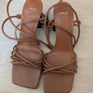 Bruna sandaler med klack från MNG - Snygga bruna sandaler från MNG med fyrkantig tå, tunna remmar och blockklack. Skorna har en trendig design med flera remmar som går över foten och runt vristen. Perfekta för att lyfta din sommarstil med en modern touch.