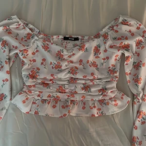 Blommig blus från Bik Bok med volang - Supersöt blommig blus från Bik Bok i vitt med rosa blommor. Toppen har långärmade puffärmar, v-ringning, rynkade detaljer framtill och en volangkant nedtill. 