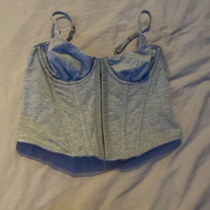 Grå korsett-topp med justerbara band - Snygg grå korsett-topp med justerbara axelband och markerade sömmar för en cool siluett. Toppen har en croppad passform och mjukt bomullsmaterial som känns skönt mot huden. Perfekt att styla med jeans eller kjol för en trendig look.