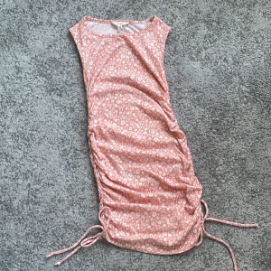 Rosa blommig kortklänning med snörning - Supersöt rosa kortklänning med vitt blommönster och snörning i sidorna. Ärmlös modell med rund halsringning och dragsko som gör att du kan justera längden. Perfekt för varma dagar och ger en fräsch och ungdomlig vibe.