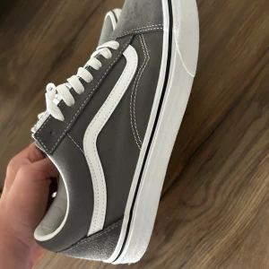 Gråa Vans Old Skool sneakers - Säljer ett par klassiska Vans Old Skool sneakers i grått med vit sula och vit sidostrip. Skorna har snörning, rund tå och är tillverkade i canvas och mocka. Perfekta för dig som gillar en clean och tidlös streetstil.