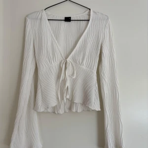 Vit omlottopp från Gina Tricot - Superfin vit topp/blis från Gina Tricot med vågigt ribbat tyg och knyt framtill. Toppen har v-ringning, långa ärmar och lätt utsvängd nederkant. Perfekt för dig som gillar en feminin och trendig look. Säljer endast då jag växt ur den! 