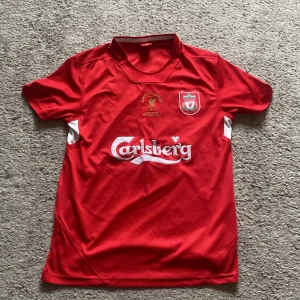 Liverpool Gerrard 2005 fotbollströja - Klassisk röd Liverpool fotbollströja med Carlsberg-tryck, klubbmärke och broderad text om Champions League-vinsten 2005 i Istanbul. Nummer 8 och Gerrard på ryggen. Tröjan har vita detaljer på sidorna och är tillverkad i lätt, ventilerande material.