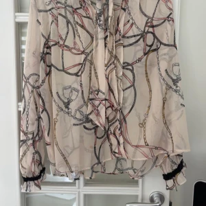 Zara blus i beige med knytning och mönster – M - Snygg blus från Zara Basic i storlek M. Tunn och luftig med kedjeinspirerat mönster i beige, rosa och svart. Har knytning fram och svarta detaljer vid krage och ärmslut. Passar både till vardag och finare tillfällen. 