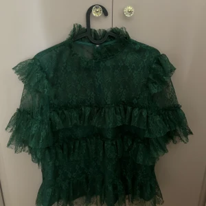 Grön volangblus i spets från By Malina - Supersnygg mörkgrön blus från By Malina med flera lager volanger och transparent spets. Blusen har hög hals och är täckt av vackra spetsdetaljer som ger en romantisk vibe. Perfekt för dig som vill sticka ut med en unik och trendig topp.