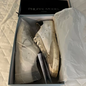 Vita Philippe Models - Snygga vita sneakers från Philippe Model Paris med svarta detaljer på hälen och grå mockadetaljer. Klassisk låg modell med snörning och rund tå. Tillverkade i skinn och mocka för en exklusiv känsla. Perfekta för dig som gillar stilrena och trendiga skor. Box och dustbag ingår. Bra skick men lite creasade och andvända