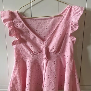 Rosa volangblus med broderi och knyt - Supersöt ljusrosa blus med volangärmar och broderade hålmönster. Toppen har v-ringning med knyt fram och är tillverkad i bomull. Perfekt för sommarens alla tillfällen när du vill känna dig extra gullig och bekväm.