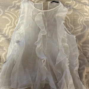 Vit volangblus från Zara - Supersöt vit blus från Zara med volangdetaljer framtill och på sidorna. Tunn och luftig modell i transparent tyg, perfekt för en fräsch och trendig look. Ärmlös design med rund halsringning.
