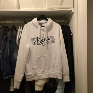 Vit hoodie  - Vit hoodie från Bear bords med svart tryck framtill och coolt surf-inspirerat motiv. Tröjan har huva med snörning och en stor känguruficka. Luvan är knappt använd och inga fläktar eller liknande!