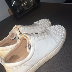 Vita Louboutin sneakers med nitar - Snygga vita sneakers från Christian Louboutin i skinn med ikonisk röd sula och coola vita nitar över tån. Klassisk snörning och låg modell. En spik är tappad och är lite sliten bak på hälen (se bild 3)