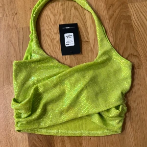 Grön glittrig topp från Fashion Nova - Säljer en grön glittrig topp från Fashion Nova med paljetter och omlott-detalj. Toppen har breda axelband och croppad passform. Storlek M. Helt ny. 