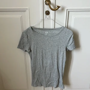 Grå ribbad topp från GAP XS - En enkel och stilren grå ribbad topp från GAP i storlek XS. T-shirten har korta ärmar och rund halsringning, perfekt för en clean och avslappnad look. Materialet är mjukt och stretchigt 