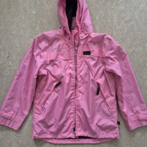 Vindjacka - Ljusrosa vindjacka med huva från Mywear Active. Jackan har dragkedja framtill, två snedställda fickor med dragkedja och justerbara ärmslut. Finns några fläckar. Perfekt tillverkad i lätt syntetmaterial, perfekt för blåsiga dagar. Snygg och sportig look med diskreta detaljer.