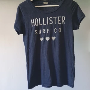 Mörkblå Hollister t-shirt med tryck - Snygg mörkblå t-shirt från Hollister med vit broderad text 'HOLLISTER SURF CO' och tre hjärtan framtill. Klassisk rund halsringning och korta ärmar. Nopprig, därav priset. Men det kan eventuellt en noppborttagare fixa (mer nopprig än vad bilderna visar, svårt att fånga på bild tyvärr) 💙