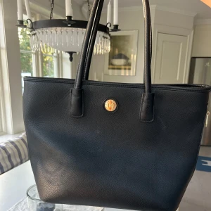 Don Donna väska - Snygg svart axelväska från don Donna i skinn med guldigt logomärke framtill. Väskan har långa handtag, dragkedja baktill och stilren, rymlig design som rymmer allt du behöver. Perfekt för dig som gillar klassisk och clean stil.