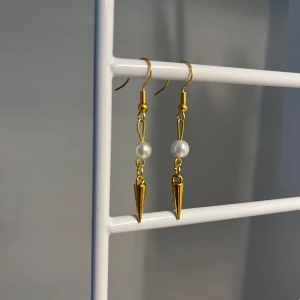 Guldiga örhängen med pärldetalj - Snygga örhängen i guldton med en vit pärla och en spetsig konformad detalj. Hängande modell som ger en cool och elegant vibe. Perfekta för att lyfta din stil med en mix av klassiskt och edgy. Handgjort 