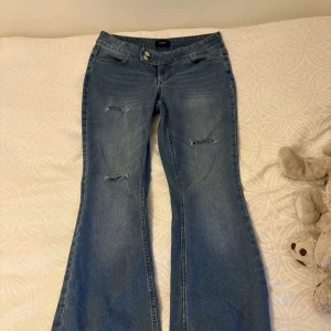 Blå bootcut jeans med slitningar - Snygga blå bootcut jeans med slitna detaljer på låren och knäna. Jeansen har klassisk femficksdesign och stängs med knapp och dragkedja. Perfekta för en avslappnad och trendig look.