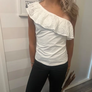 Vit one shoulder topp med volang - Superfin vit topp i bomull med bred broderad volang över ena axeln. One shoulder-modell som ger en snygg och trendig look. Perfekt till jeans eller kjol när du vill sticka ut lite extra.