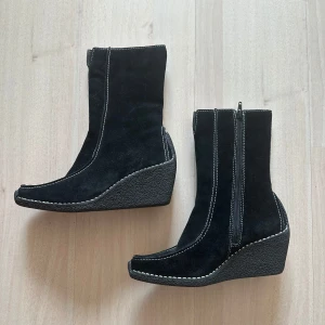 Svarta mockastövletter - Snygga svarta stövletter i mocka med markerade vita sömmar och dragkedja på sidan. Skorna har spetsig tå och en rejäl kilklack som ger extra höjd och cool siluett. 