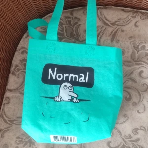 Turkos shoppingväska från Normal - Turkos shoppingväska i syntetmaterial med dubbla handtag. På framsidan finns ett tryck med texten 'Normal' och en tecknad figur. Väskan är rymlig och lätt att bära med sig till skolan, stan eller när du handlar. Återanvändbar och med cool, enkel design. Är perfekt ifall man vill ha en påse till saker man har köpt istället för att köpa en påse på affären 