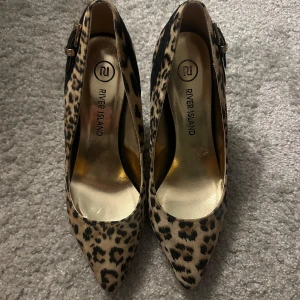 Leopardmönstrade pumps från River Island - Snygga pumps från River Island i leopardmönster med spetsig tå med klack. Skorna har svart band och guldfärgat spänne vid hälen. Utsidan är i mockaliknande material som ger en lyxig känsla.