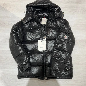 Svart Moncler pufferjacka med huva - Säljer en svart pufferjacka från Moncler med glansig finish, huva och dragkedja framtill. Jackan har Moncler-logga på ärmen, praktiska fickor och coola detaljer som elastiska muddar. Perfekt för dig som vill ha en varm och snygg vinterjacka med streetkänsla.